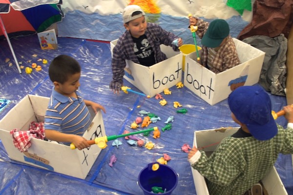 Children sitting inside boxes trying to pick up items on the floor with a long toy.